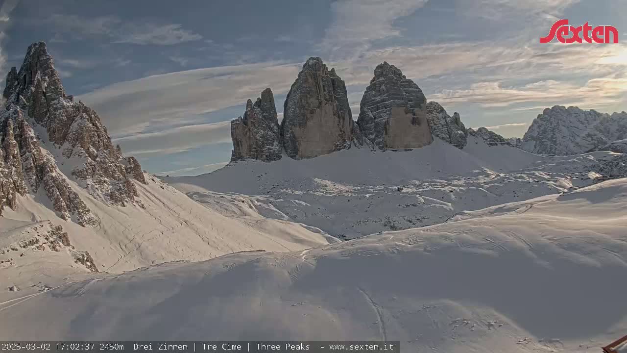 A snow-covered mountain range with prominent, tall, rocky peaks is visible under a partly cloudy sky.
