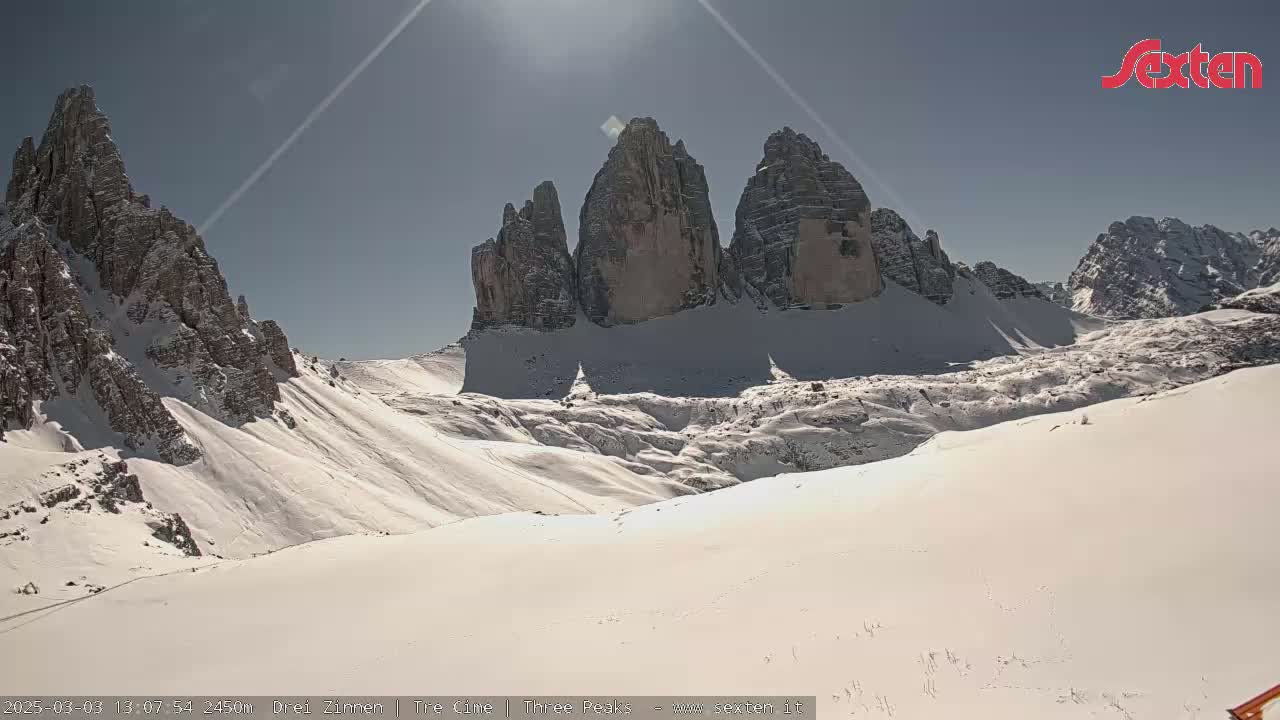 A sunny, snow-covered mountain valley features three prominent rock formations in the background.