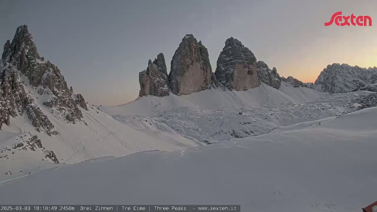 A snow-covered mountain range, illuminated by the soft light of sunrise or sunset, features several prominent, sharply peaked rock formations.