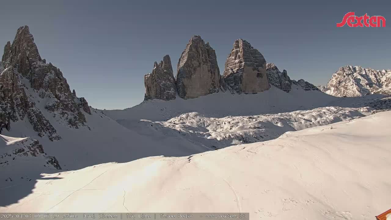 A snow-covered mountain range with several prominent peaks, under a clear, sunny sky.