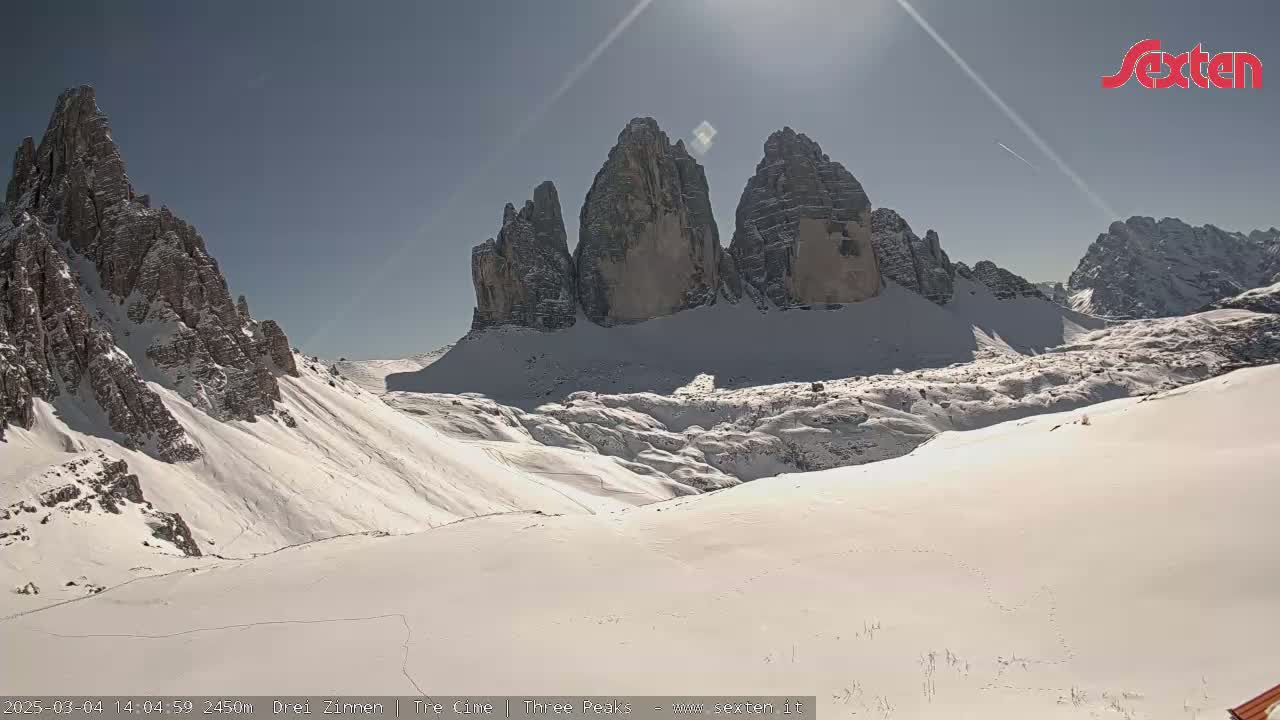 A sunny day reveals snow-covered mountains, with a prominent group of three tall, slender peaks in the center.