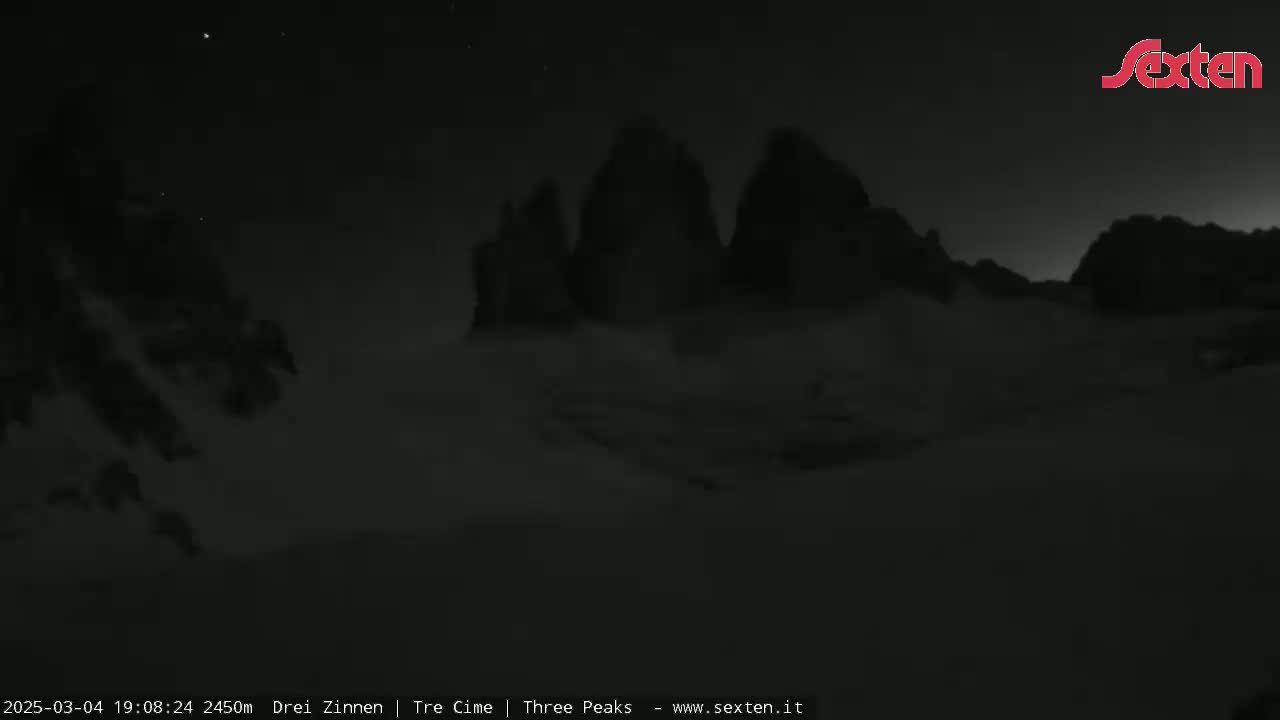A nighttime, low-light view shows three dark, mountain peaks covered in snow under a clear, starry sky.