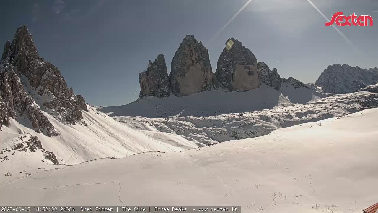 A sunny, snow-covered mountain valley is viewed from a high vantage point, showing several prominent rock formations.