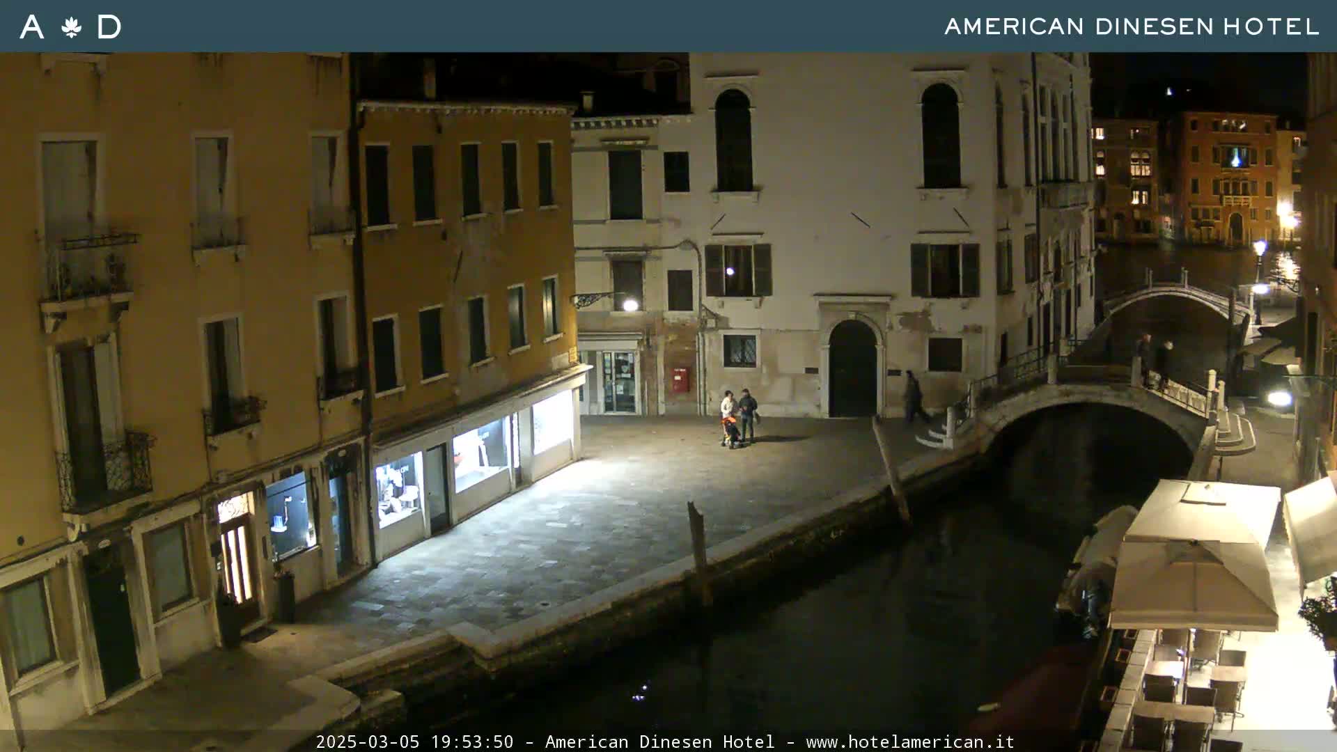 A nighttime view of a canal in Venice, showing several buildings and a small group of people, with a bridge and a restaurant visible in the distance.