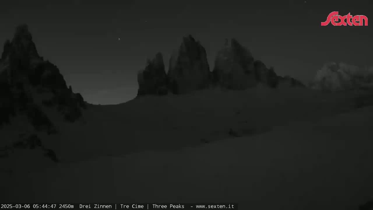 A nighttime, low-light image shows a range of dark, snow-covered mountains under a mostly clear sky with a few visible stars.