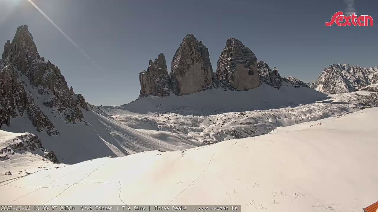 A sunny, snow-covered mountain landscape features a prominent rock formation of three tall peaks.