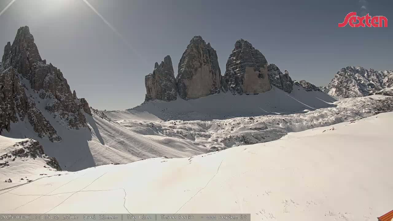 A sunny, snow-covered mountain landscape features a prominent group of three jagged peaks in the background.
