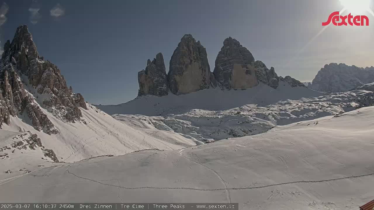 A sunny, snow-covered mountain valley with prominent, jagged peaks in the background.