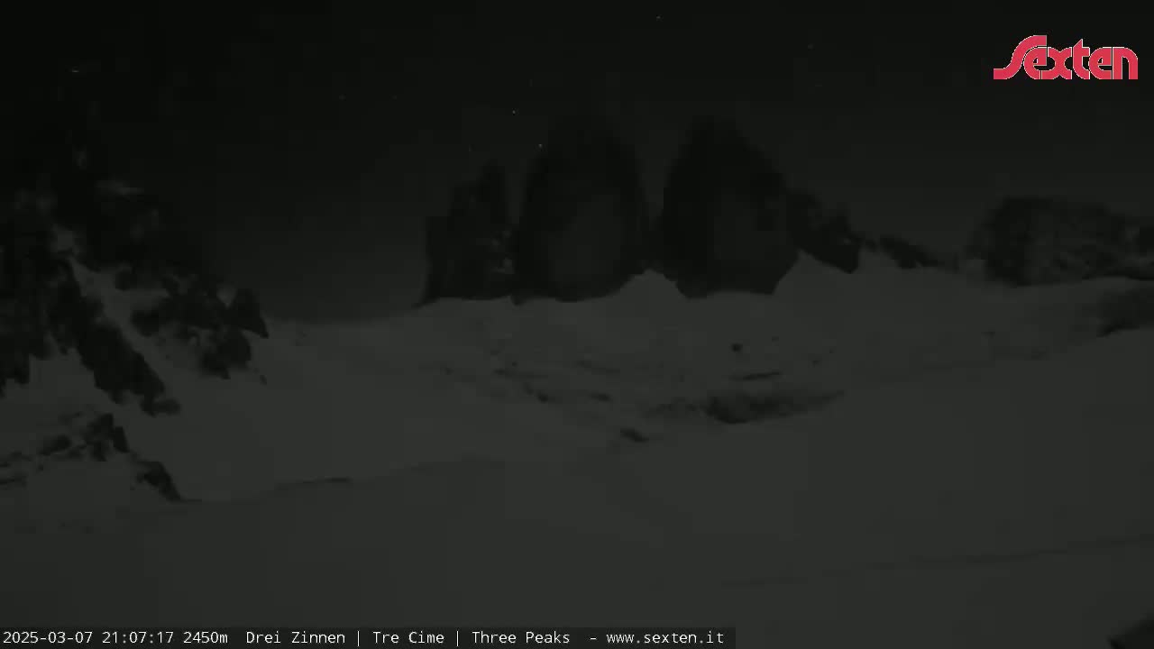 A nighttime, snow-covered landscape features dark, mountainous silhouettes under a mostly clear sky.