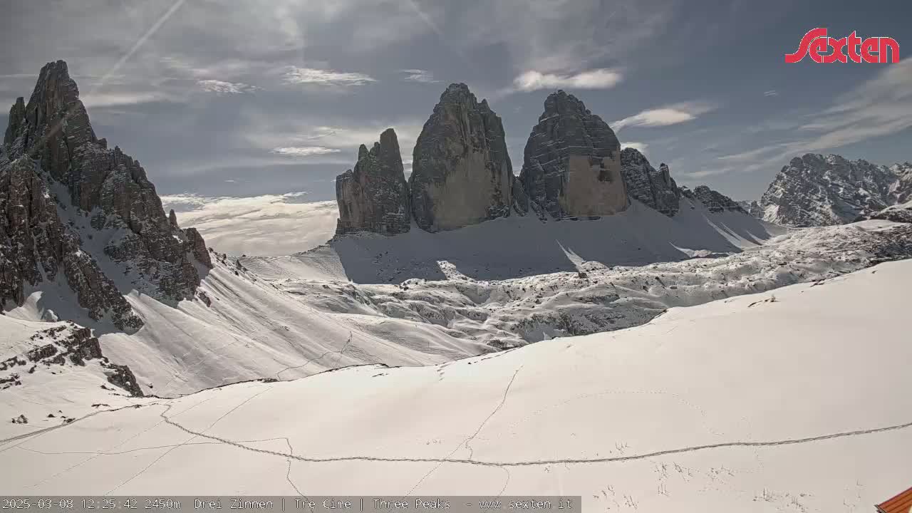 A snow-covered mountain landscape features a prominent rock formation of several tall, slender peaks under a partly cloudy sky.