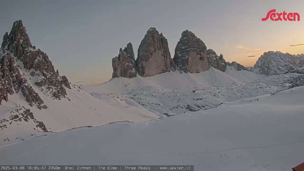 A snow-covered mountain range with prominent rock formations is shown at sunset.