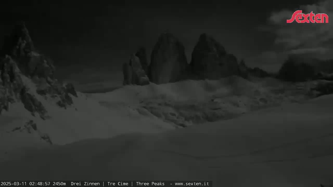 A snow-covered landscape at night features a range of dark, jagged mountains under a cloudy sky.