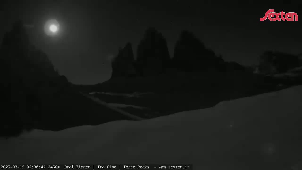 A bright moon illuminates snow-covered ground in the foreground with dark, mountainous silhouettes in the background under a clear night sky.