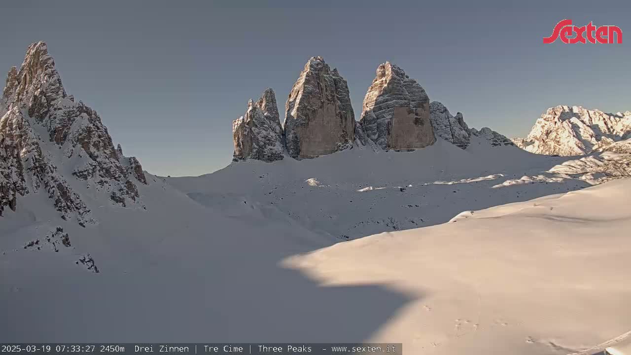 A snow-covered mountain range with several prominent peaks is seen under a clear, bright sky.