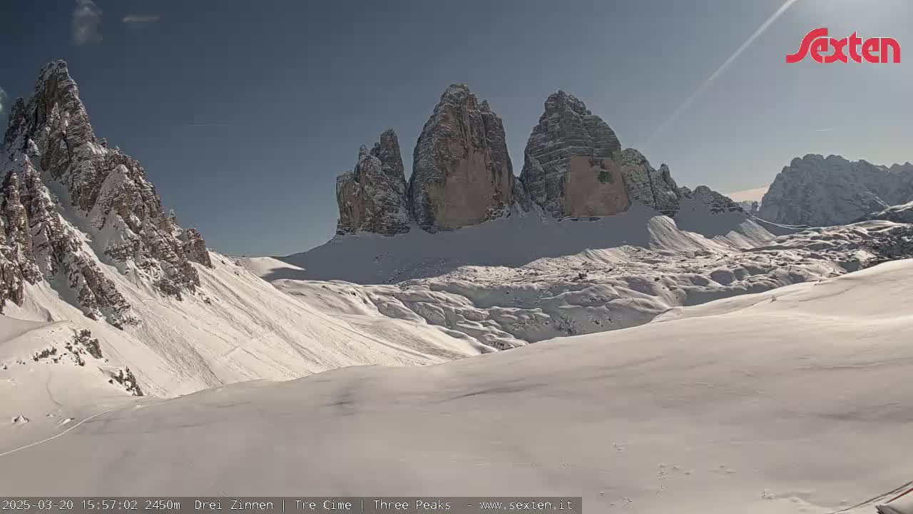 A sunny, snow-covered mountain landscape features three prominent, jagged peaks in the background.