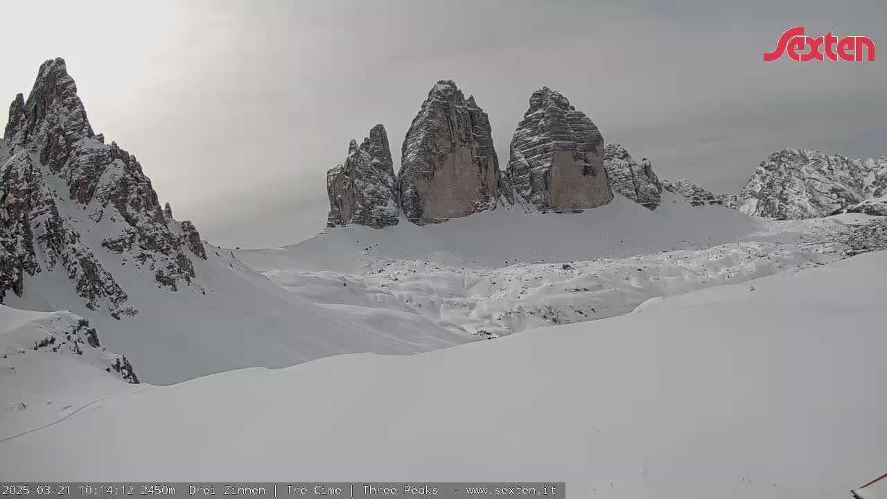 A snow-covered landscape features several tall, rocky peaks under an overcast sky.