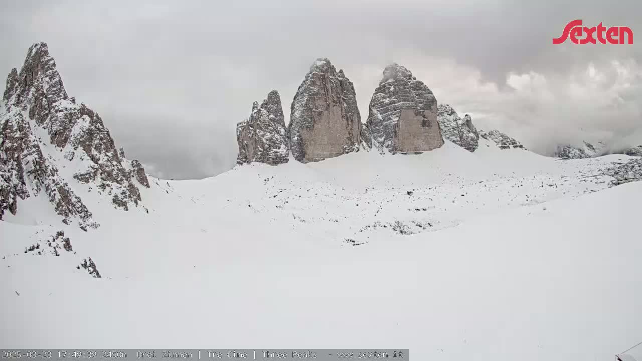 A snow-covered landscape features a prominent group of three tall, rocky peaks under an overcast sky.