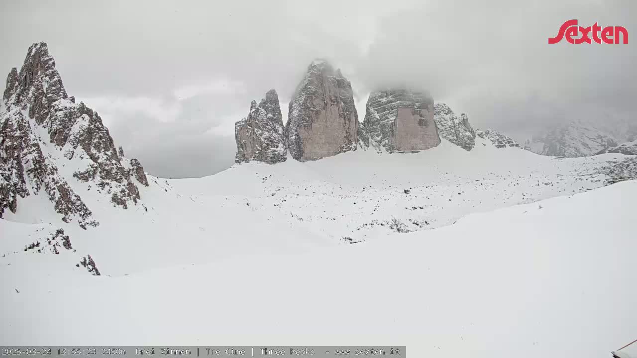 Snow-covered mountains, including three prominent peaks partially obscured by clouds, are visible under an overcast sky.
