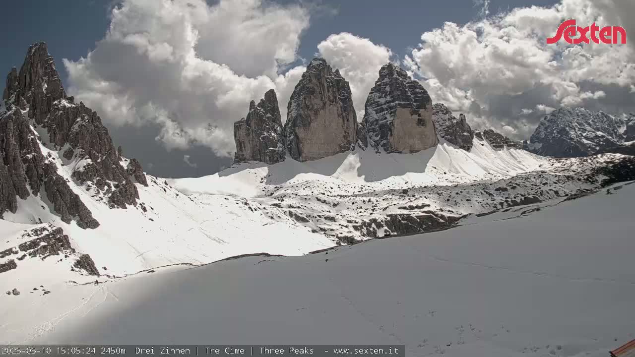 A snow-covered mountain landscape with three prominent rock peaks under a partly cloudy sky.