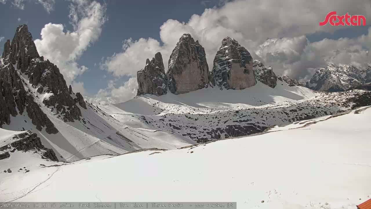 A snow-covered mountain range, partially cloudy sky, with several prominent, sharply peaked rock formations visible in the mid-ground.