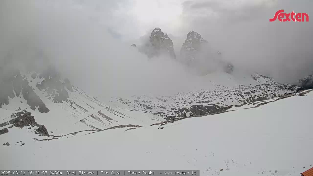A snow-covered mountain valley is shrouded in fog, with rocky peaks partially visible in the distance.