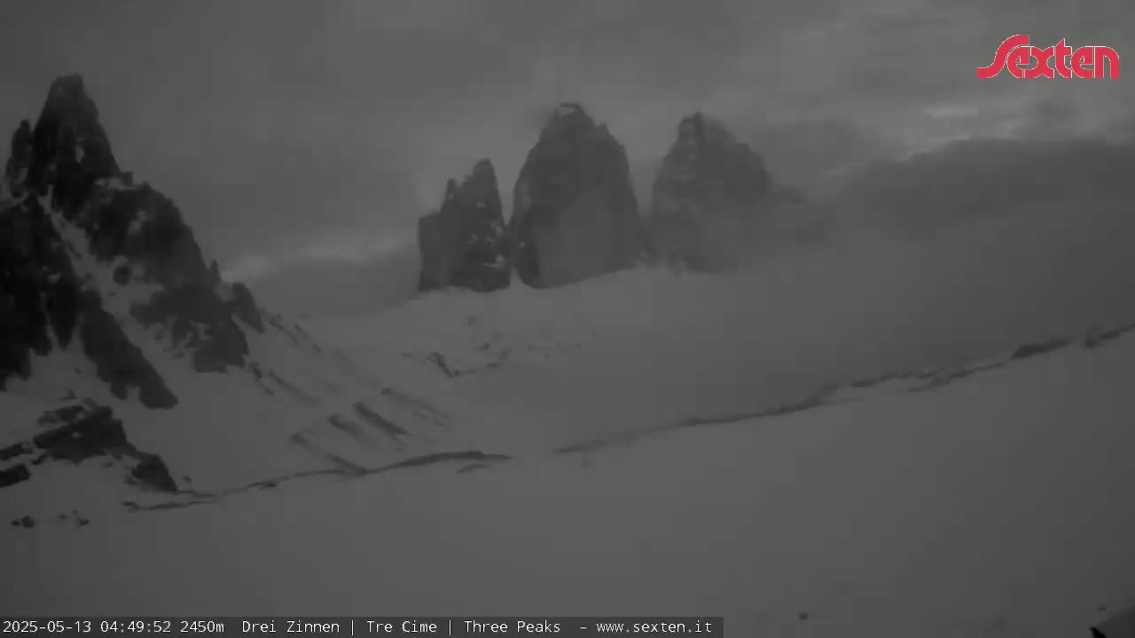 A snowy, overcast mountain landscape shows three prominent peaks partially obscured by fog or clouds.