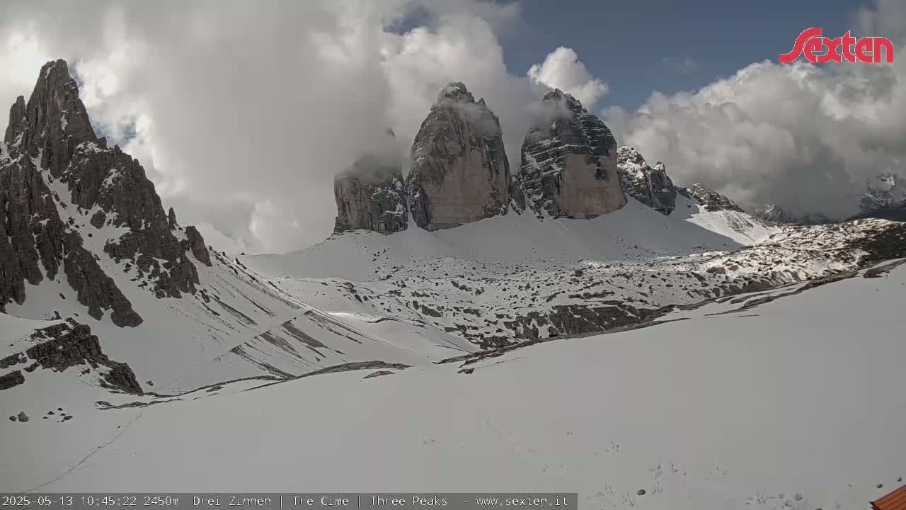 A snow-covered mountain valley with three prominent rock formations partially obscured by clouds under partly sunny skies.