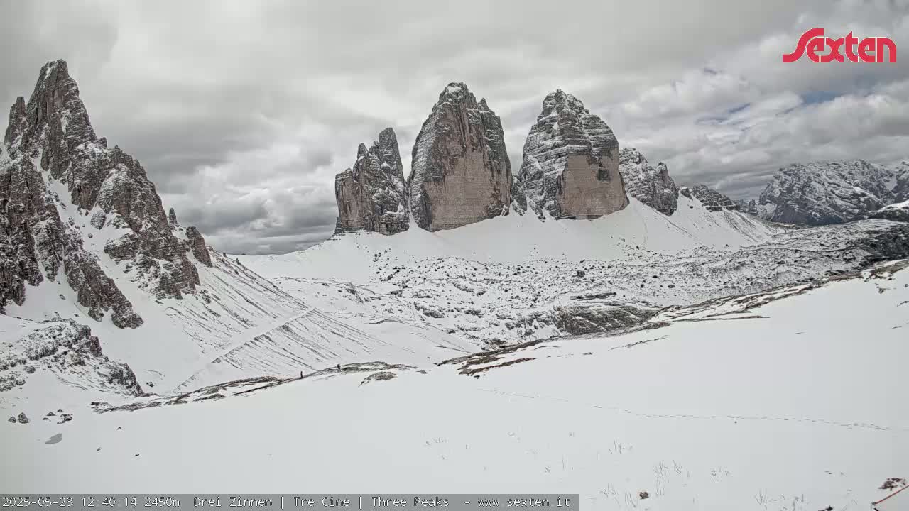 A snow-covered valley with prominent, jagged, snow-capped peaks under a cloudy sky.
