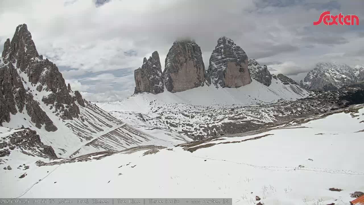 A snow-covered mountain valley with three prominent peaks under a cloudy sky.