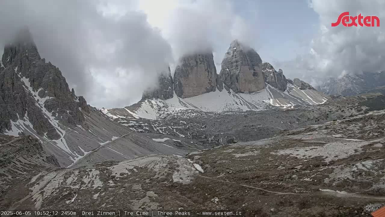 A mountain range with snow-capped peaks partially obscured by clouds is viewed from a lower elevation.