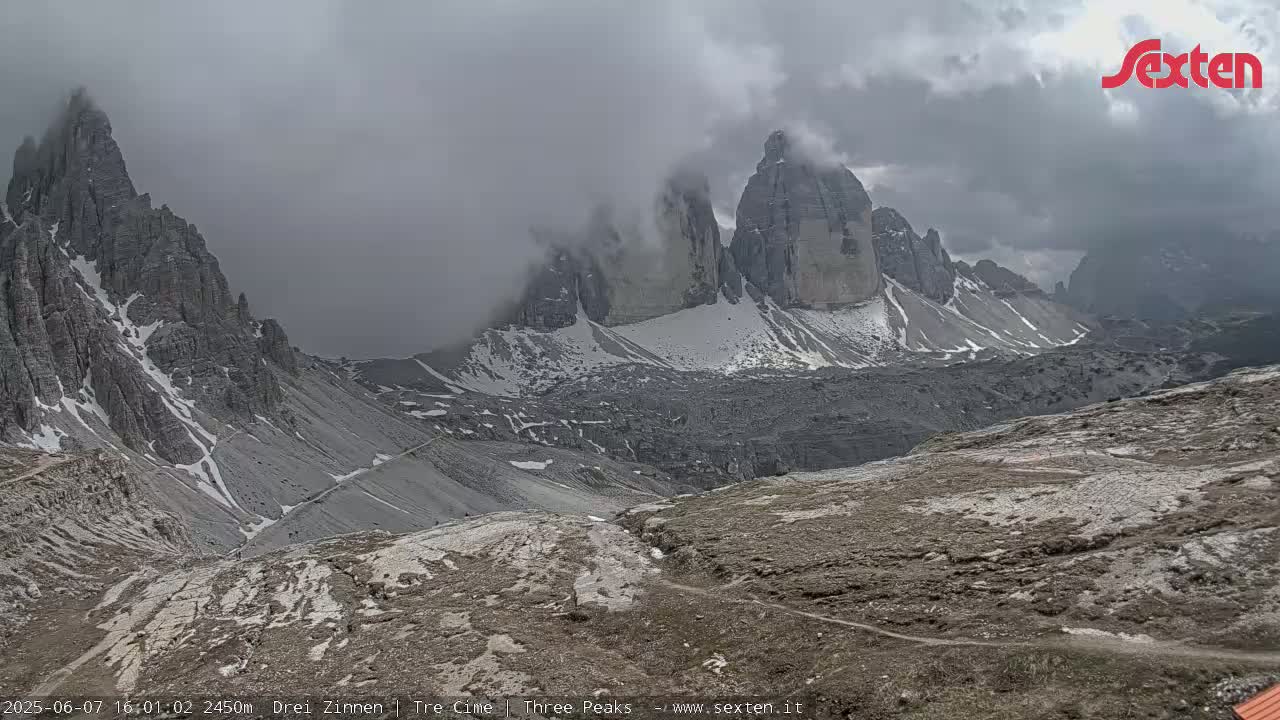 A mountain range with snow-capped peaks partially obscured by clouds and mist is viewed from a high vantage point overlooking a rocky, barren landscape.