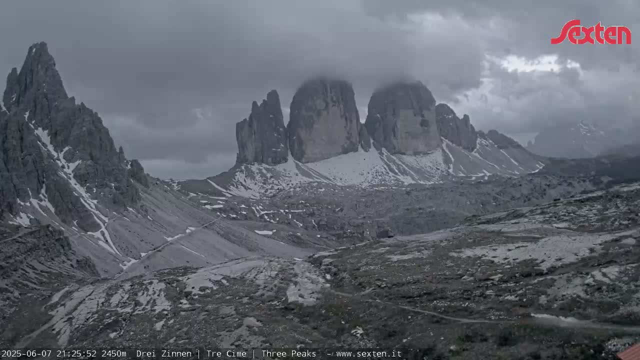 A mountainous landscape with snow-capped peaks partially obscured by clouds under an overcast sky.
