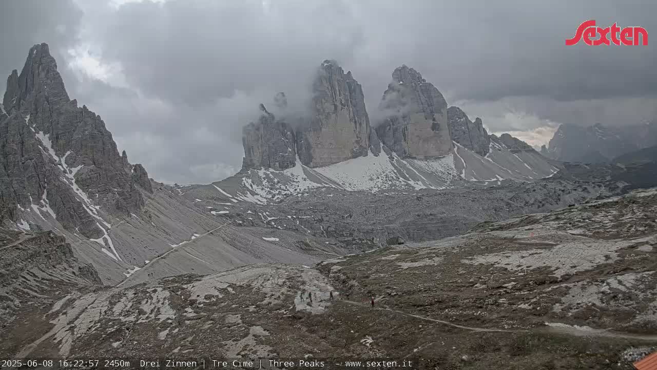 A mountain valley with several tall, grey, snow-dusted peaks partially obscured by clouds under an overcast sky.