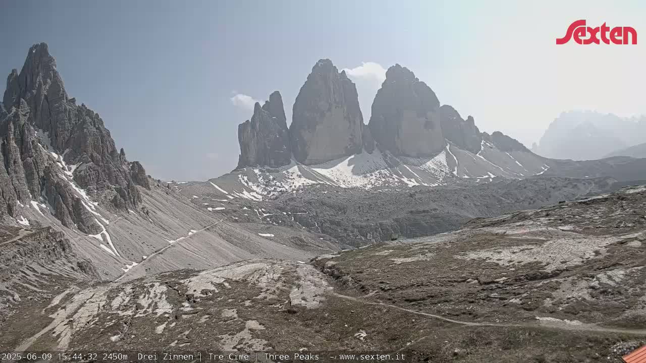 A hazy, sunny day reveals a view of several tall, gray, rocky mountains with patches of snow, overlooking a mountainous, barren landscape.