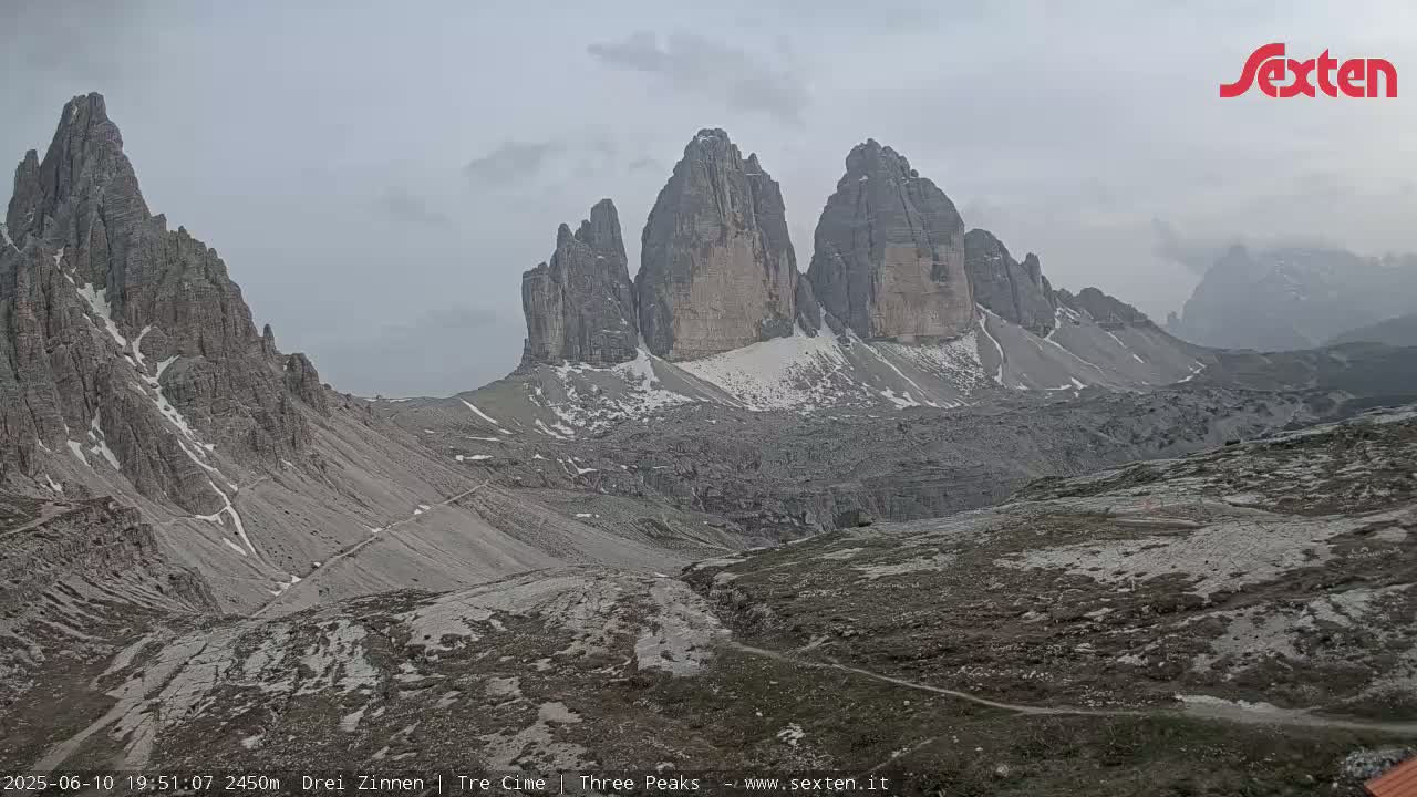 A mountain range with several tall, rocky peaks partially covered in snow is visible under an overcast sky.