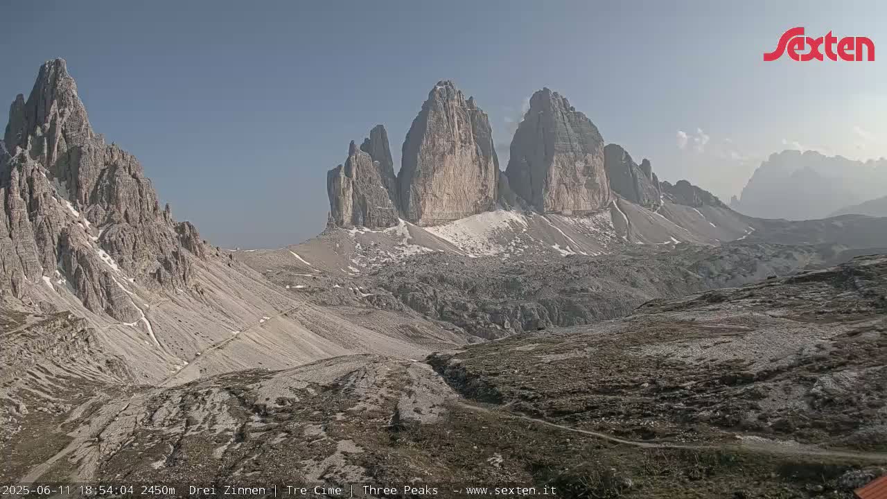 A rocky, mountainous landscape with a few patches of snow under a clear, light blue sky shows several tall, jagged peaks in the background.