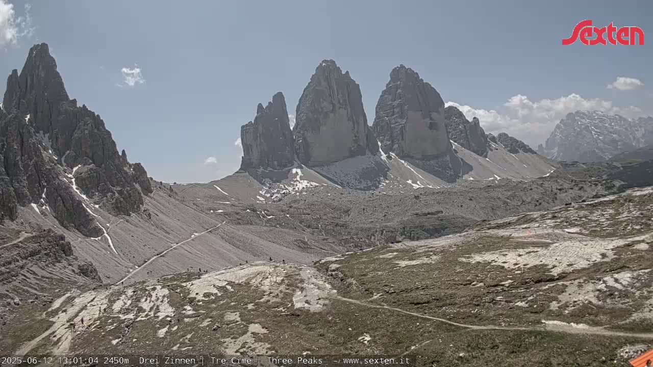 A mostly sunny day reveals a rocky, mountainous landscape with several tall, jagged peaks and winding trails, some patches of snow visible on higher elevations.