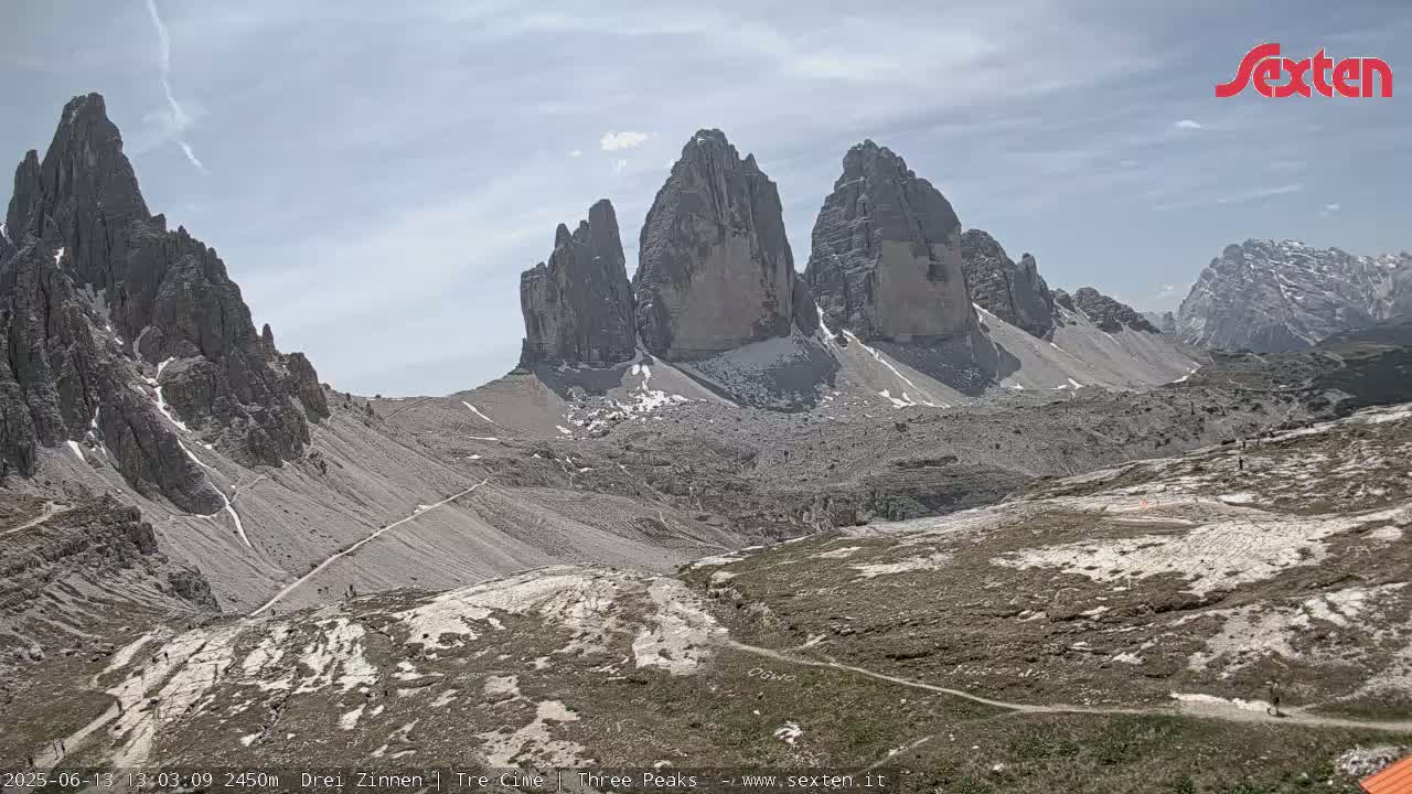A high-angle view shows three prominent, tall, rocky peaks under a mostly clear sky with a few wispy clouds, and a valley below with sparse vegetation and a few people visible on trails.