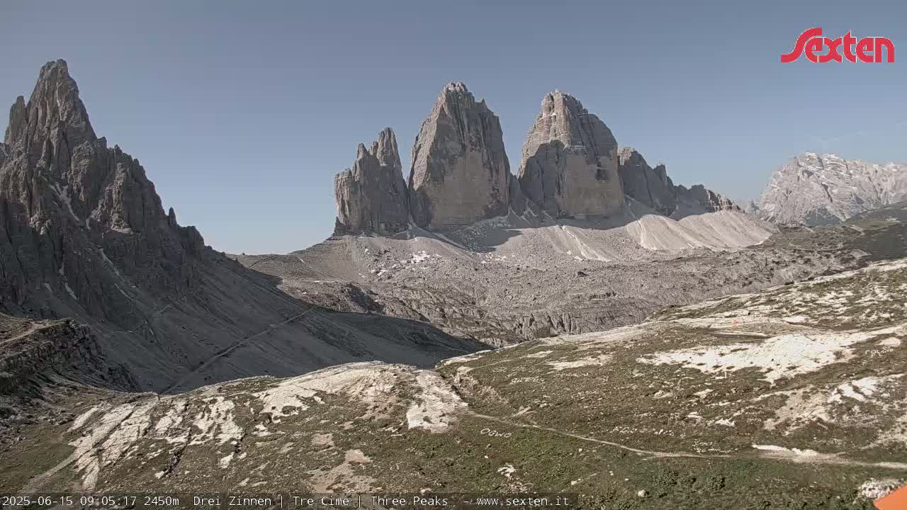 A high-angle view on a sunny day shows a rocky, mountainous landscape with a prominent group of three tall, slender peaks and winding paths.