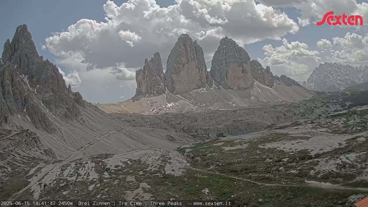 A partly cloudy day reveals a mountainous landscape featuring a prominent rock formation of three tall peaks, with a valley and trails visible in the foreground.