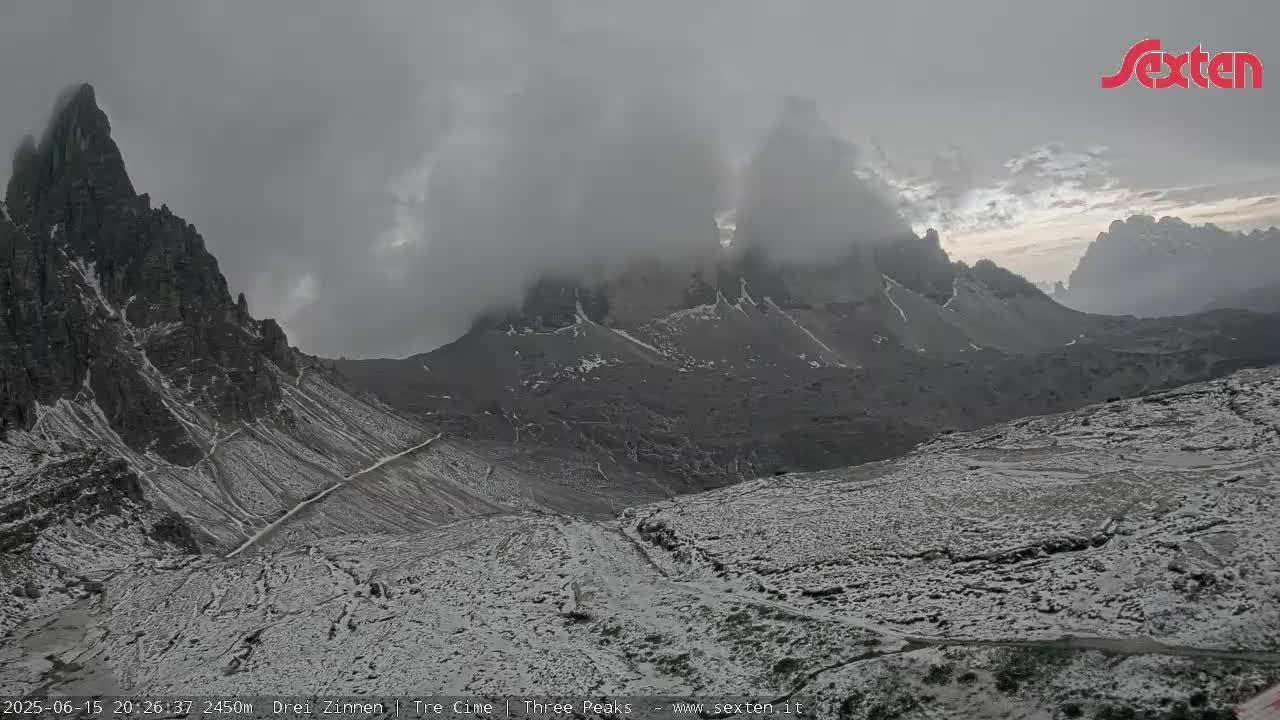 A snow-dusted mountain landscape with dark, jagged peaks partially obscured by low-lying clouds under an overcast sky.