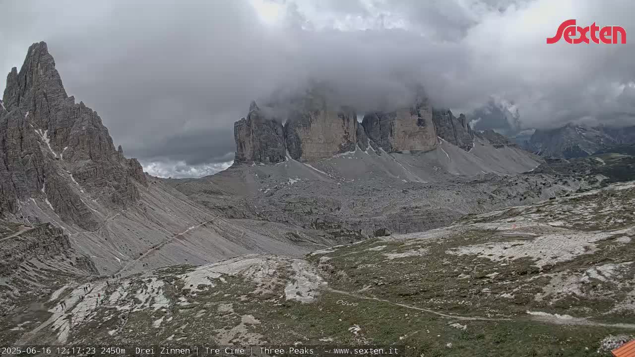 A mountain range partially obscured by clouds is seen from a high vantage point, showing rocky terrain and winding paths below.