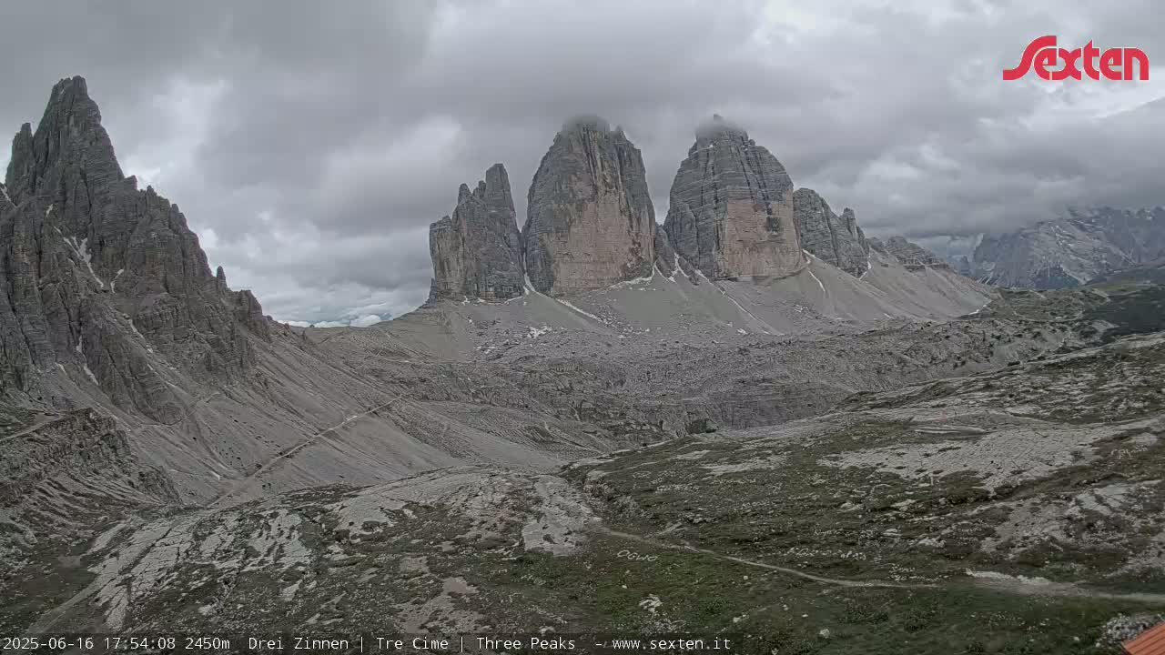 A mountainous landscape under a cloudy sky shows several rocky peaks and a valley with sparse vegetation.