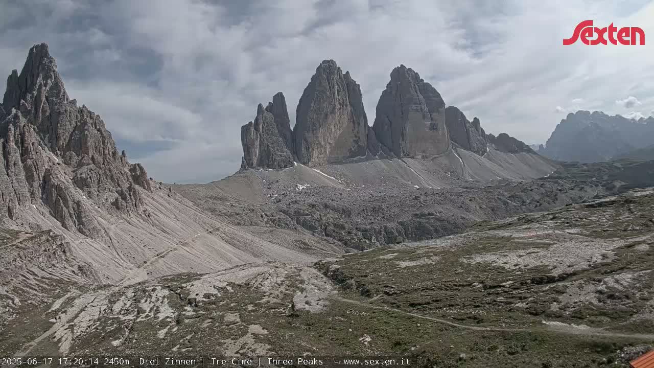 A mountainous landscape under a cloudy sky shows several rocky peaks and a valley with sparse vegetation.