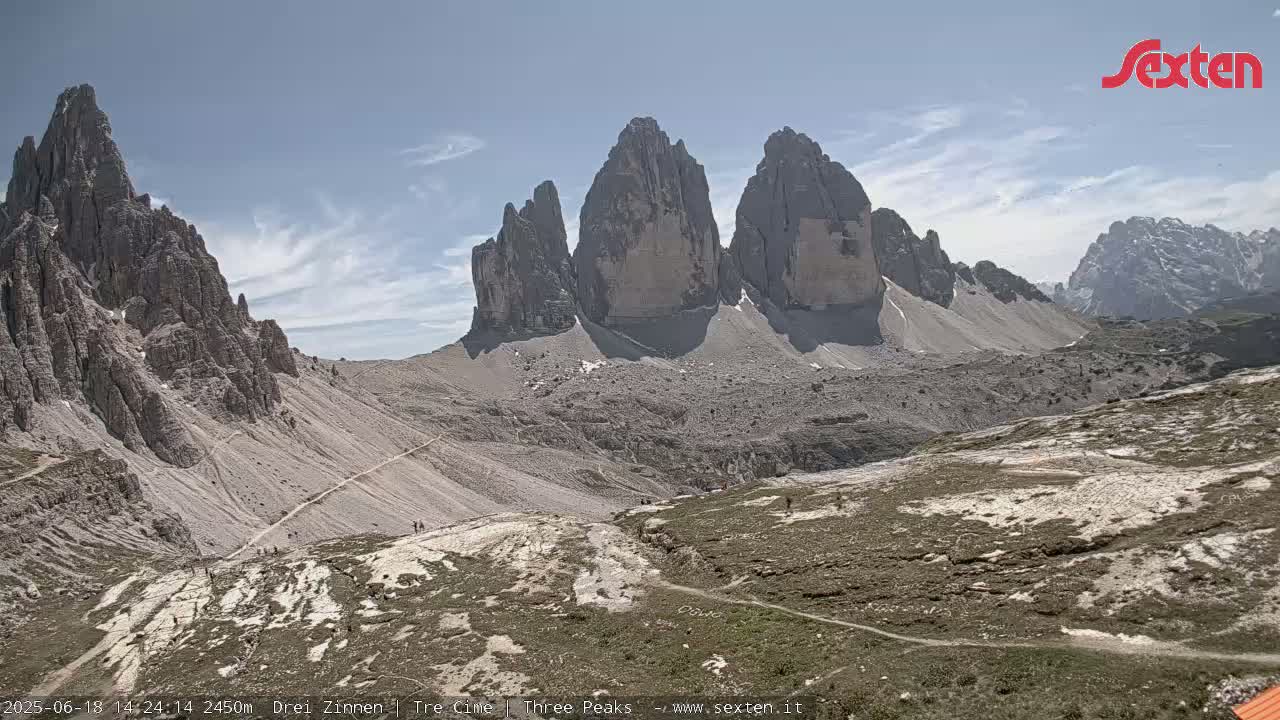 A high-angle view shows several large, rocky, gray mountains under a mostly sunny sky, with some sparse vegetation and trails visible in the foreground.