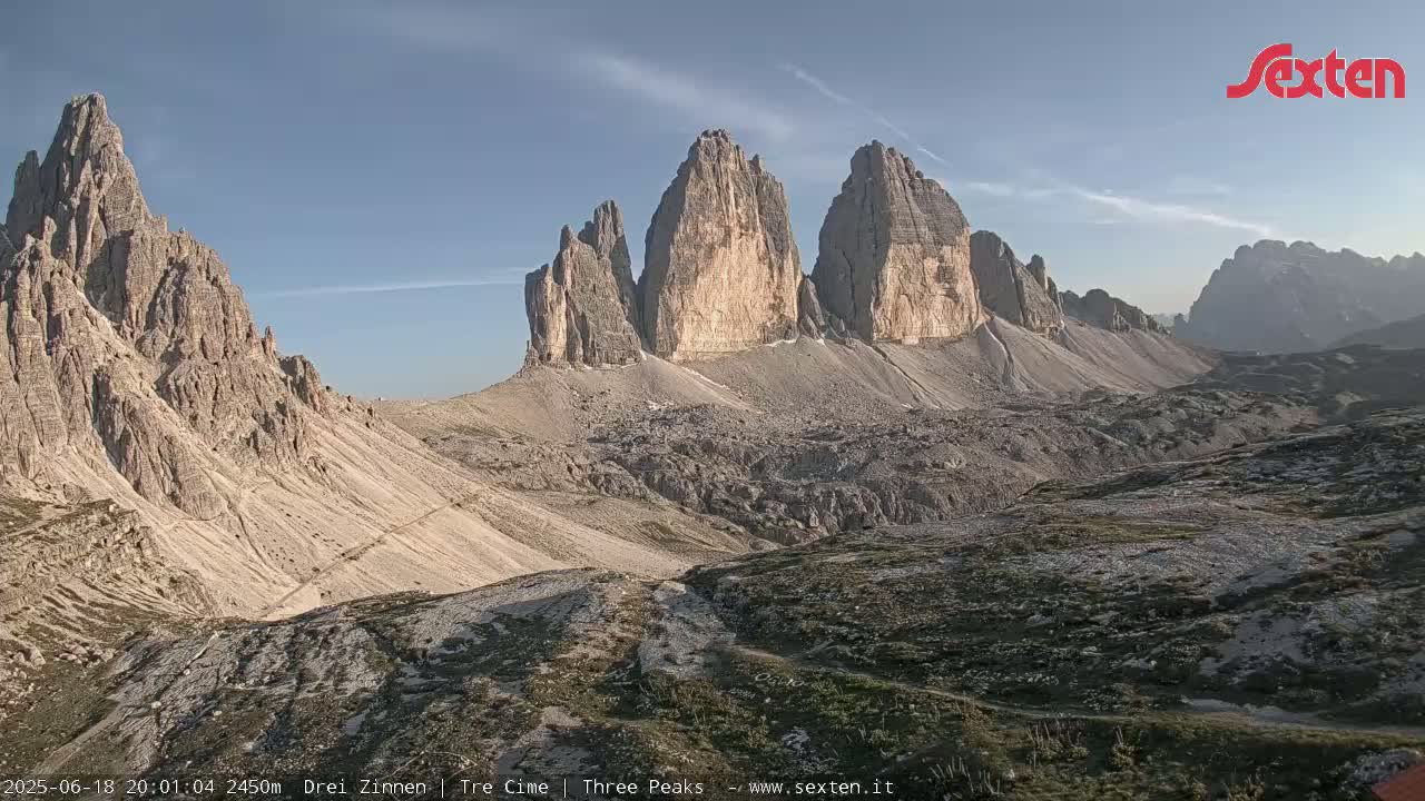 A panoramic view of a rocky mountain range under a clear, bright sky shows several tall, jagged peaks.