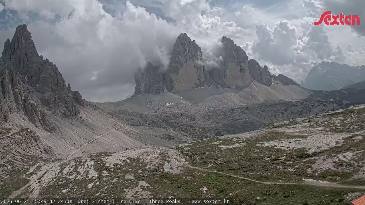 A mountain valley with rocky terrain and a few trails is partially obscured by clouds under a partly cloudy sky.