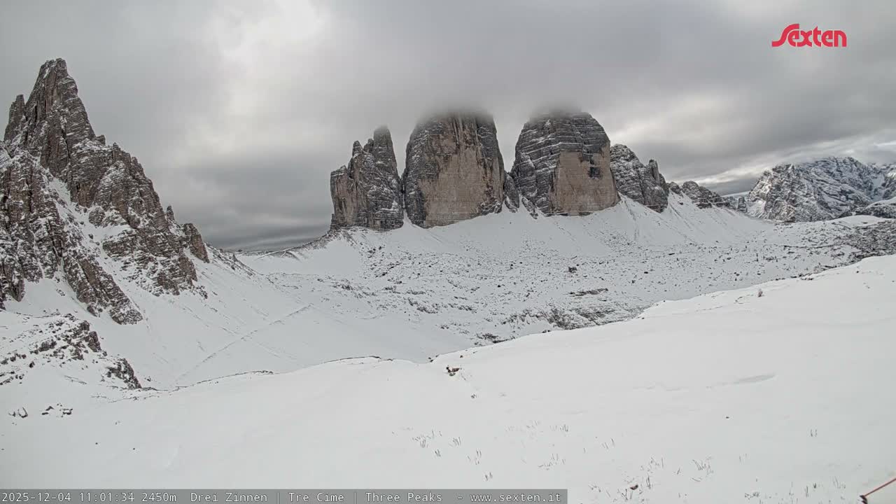 A vast, snow-covered mountain landscape features several rugged, rocky peaks, with the tallest ones partially obscured by low, heavy clouds under an overcast sky.