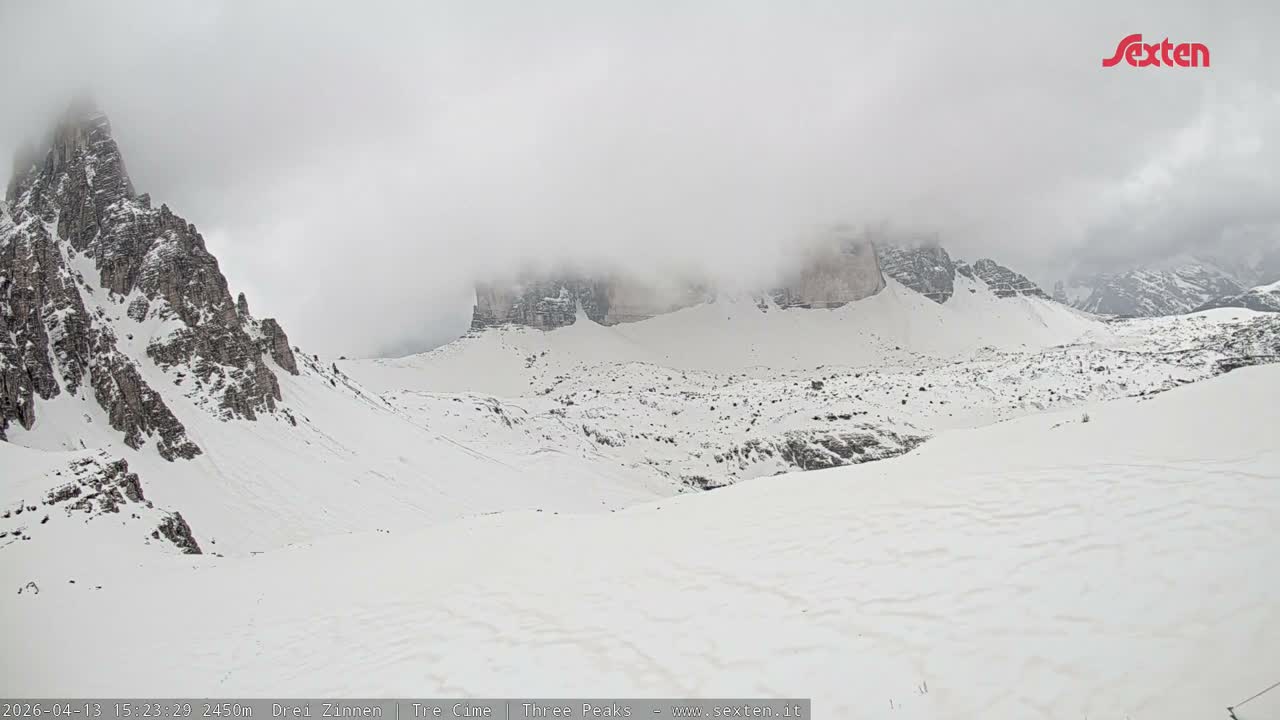 A vast, snow-covered mountain landscape features several rugged, rocky peaks, with the tallest ones partially obscured by low, heavy clouds under an overcast sky.
