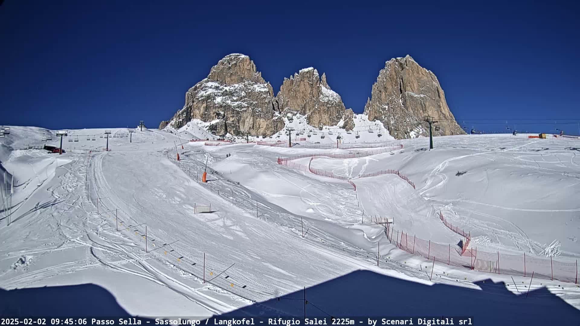 A snow-covered ski slope with ski lifts and several rocky peaks in the background under a clear blue sky.